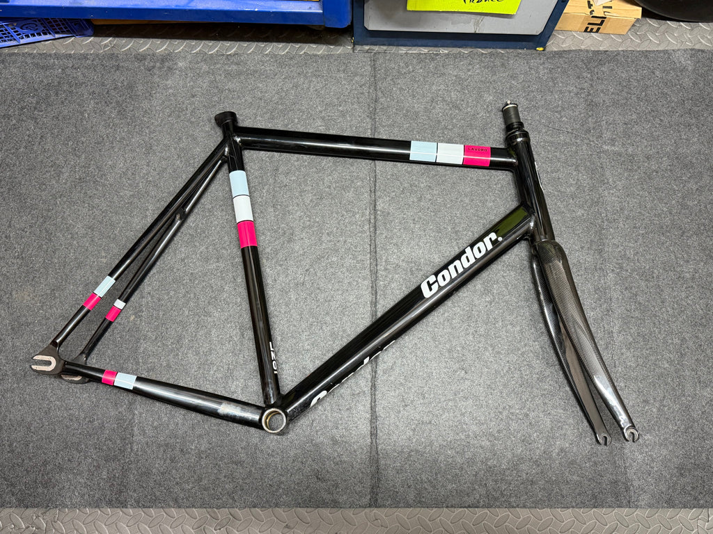 A black Condor bicycle frame laid flat on a gray textured floor, with white, pink, and light blue accent strips on the top tube and seat tube.
